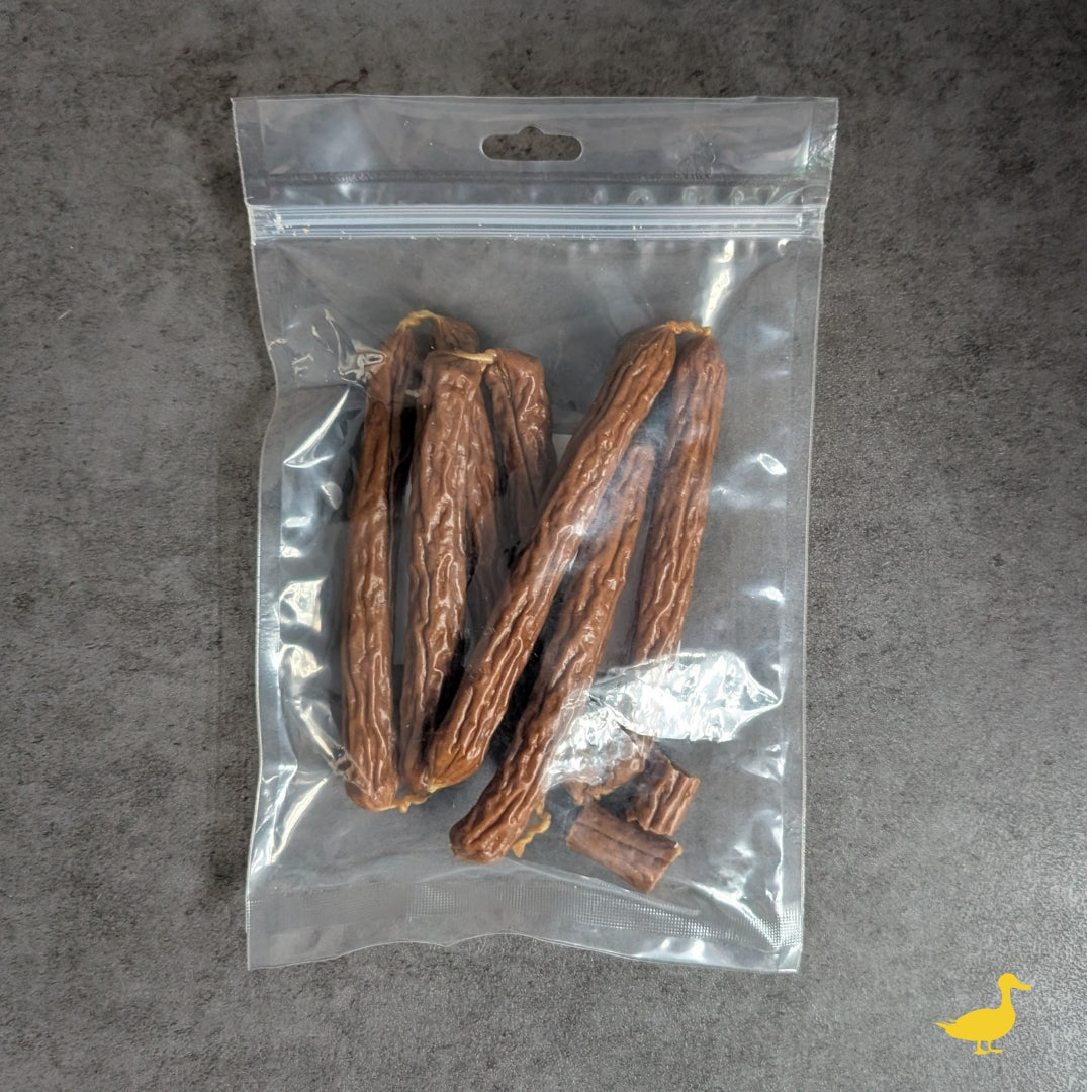 Transparenter Zip-Beutel mit braunen Enten Kabanossi-Sticks (~15 cm), ein Stück gebrochen, grauer Untergrund