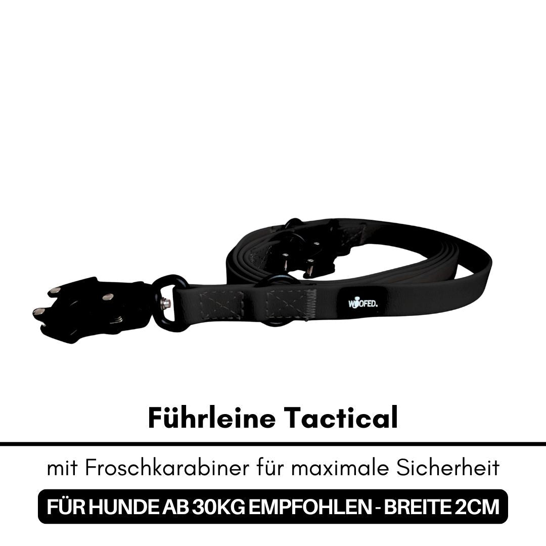 Schwarze taktische Führleine aufgewickelt mit Froschkarabiner, D-Ring und WOOFED-Label, Vorderansicht auf weißem Hintergrund