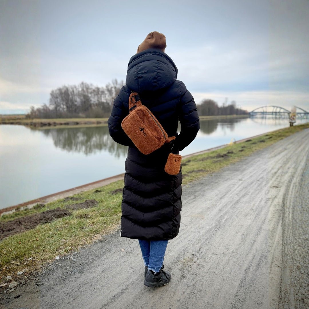 Person von hinten mit brauner Teddy-Gassitasche quer über dem Rücken und kleinem Beutel am Gurt, Spazierweg am Fluss