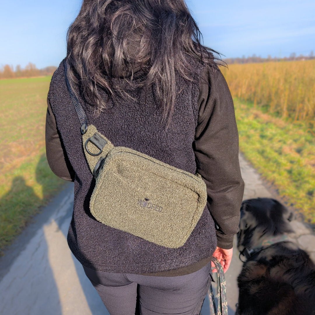 Olivgrüne Teddy Gassitasche quer getragen, rechteckig mit Reißverschluss und D-Ring, Rückenansicht auf Feldweg neben Hund