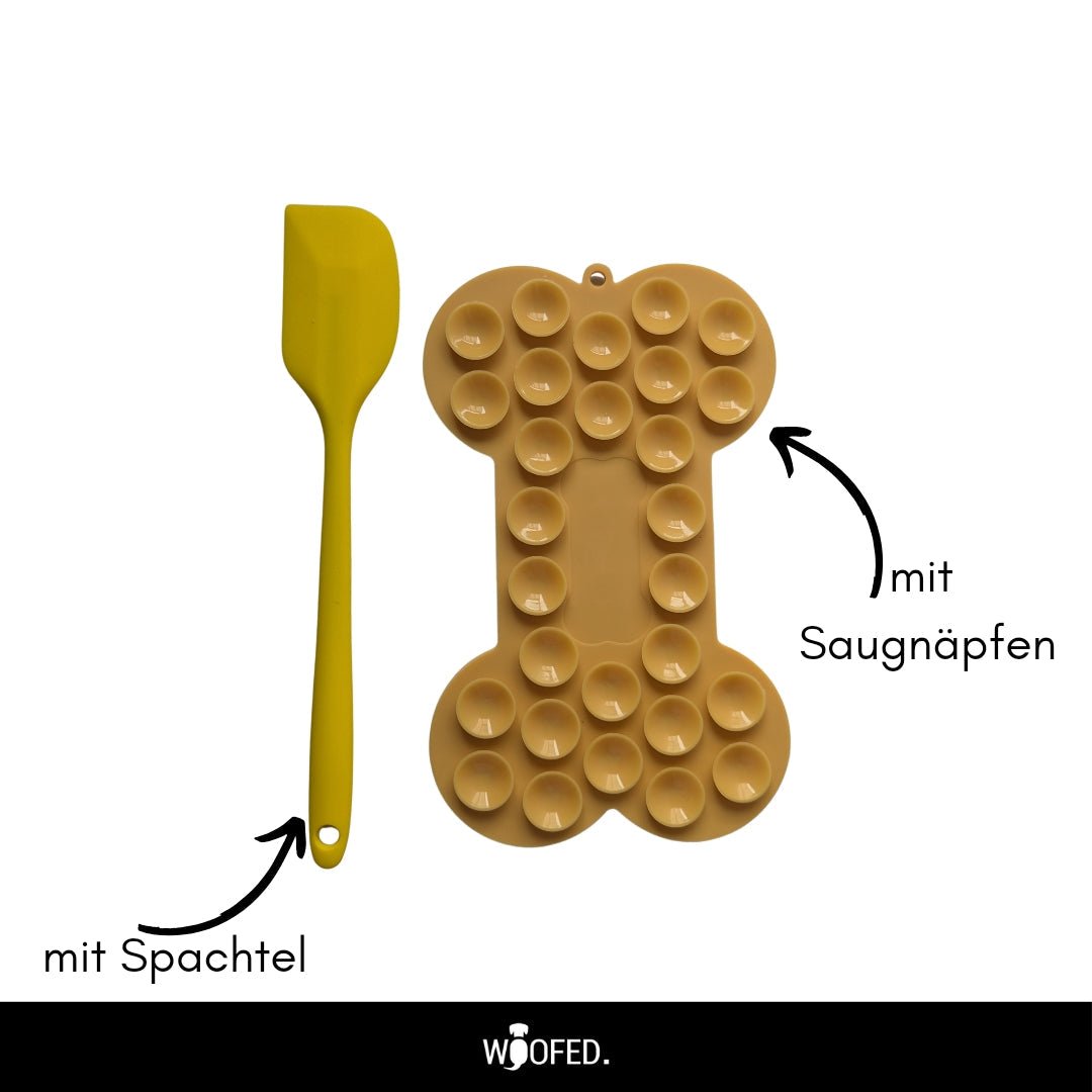 Knochenförmige, beige Schleckmatte mit runden Saugnäpfen, Draufsicht; gelbe Spachtel links