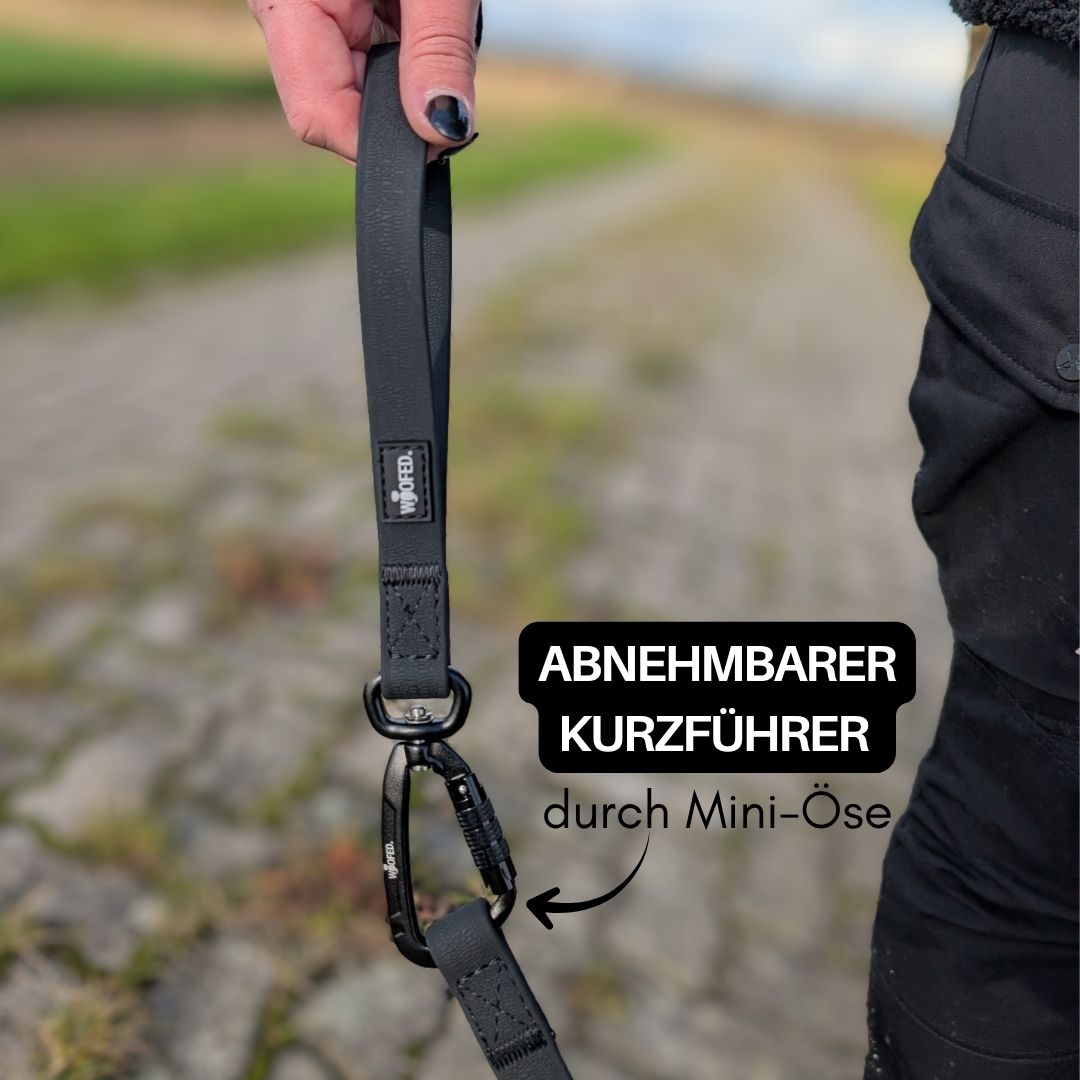 Hand hält schwarzen Kurzführer mit Karabiner und WOOFLEX-Label, Nahaufnahme vor gepflastertem Weg