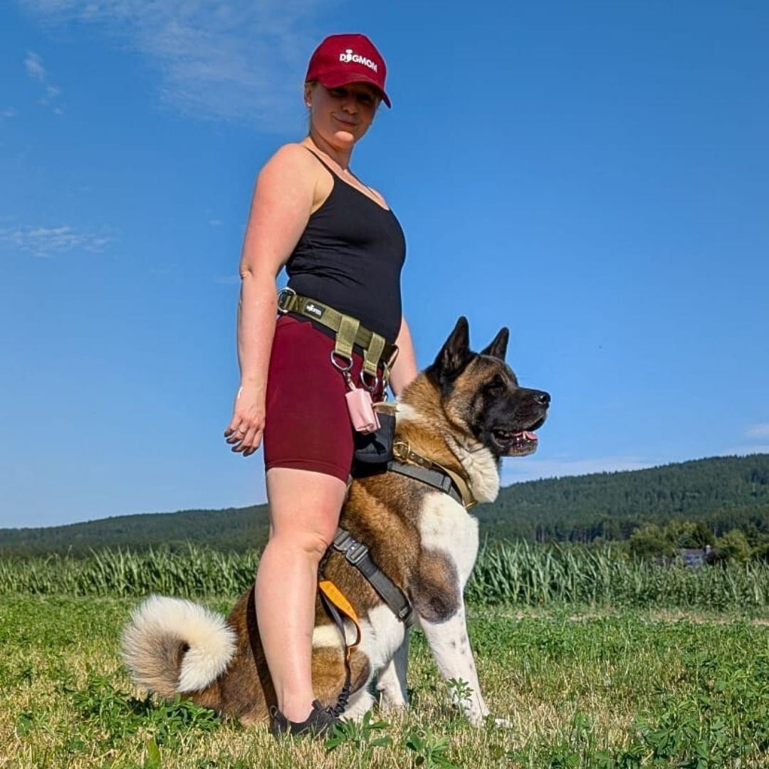 Frau seitlich mit olivgrünem Gassigürtel, taktischer Schnalle, D‑Ring und pinkem Beutel, daneben sitzender Hund