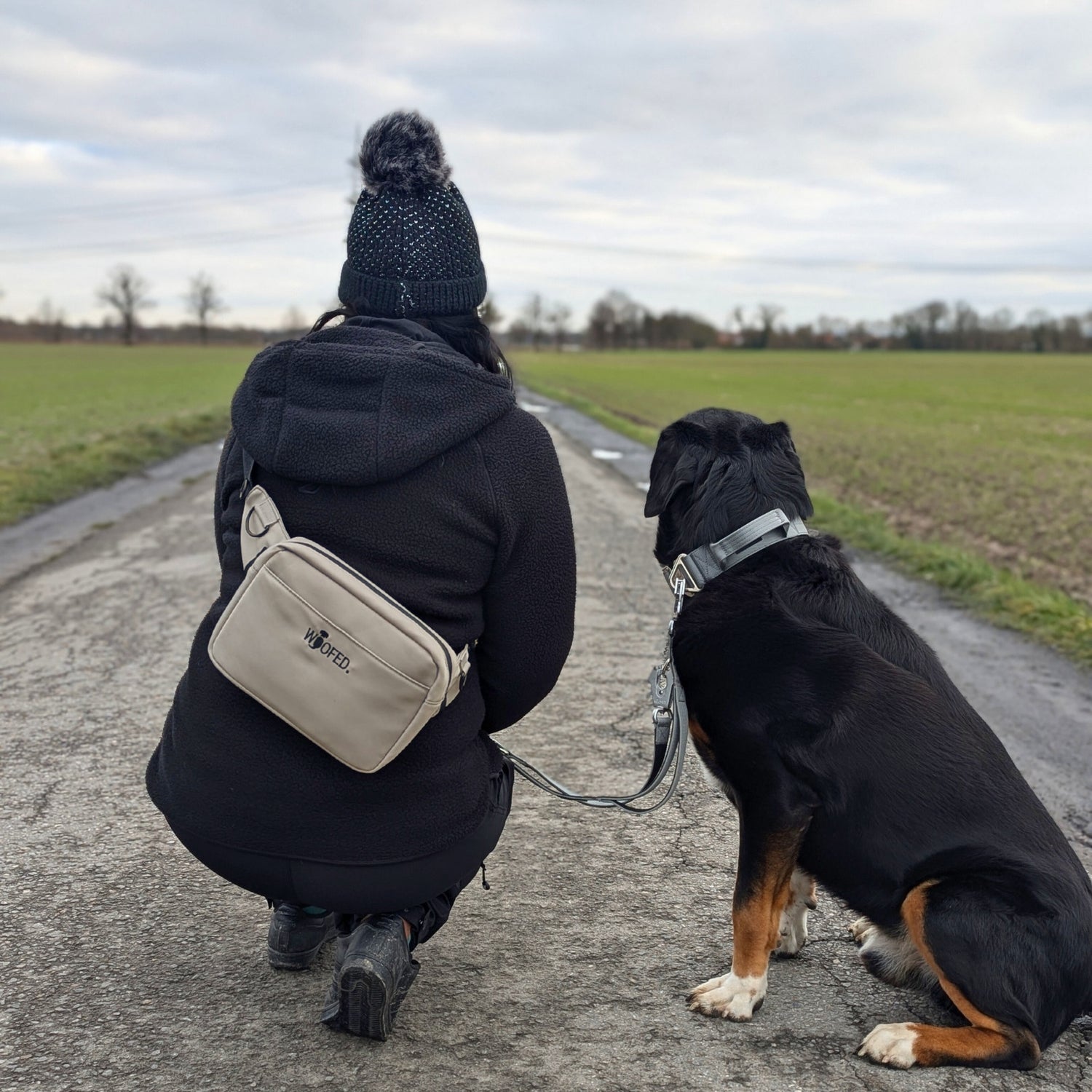 Rückenansicht: beige Gassitasche quer getragen mit WOOFED-Logo, kniende Person neben schwarzem Hund auf Feldweg