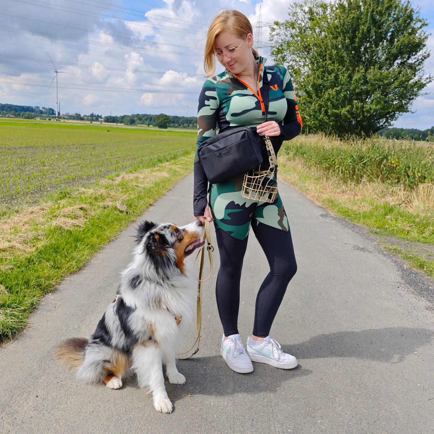 Schwarze Gassitasche quer über der Hüfte getragen, Vorderansicht; Frau mit Hund und Leine auf ländlichem Spazierweg