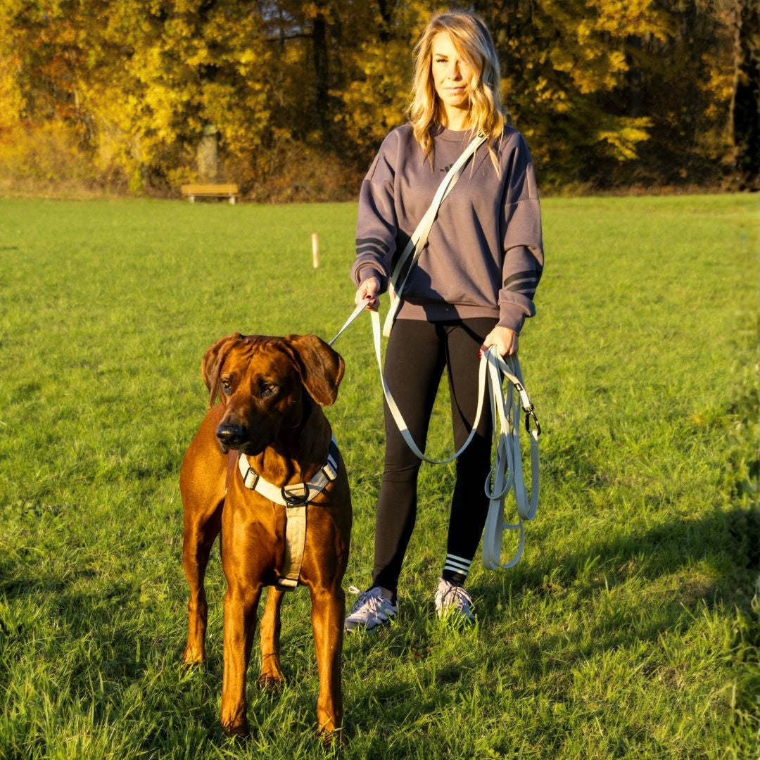 Frau hält beige WOOFLEX Desert Sand Schleppleine über der Schulter, in der Hand und am braunen Hundegeschirr auf Wiese
