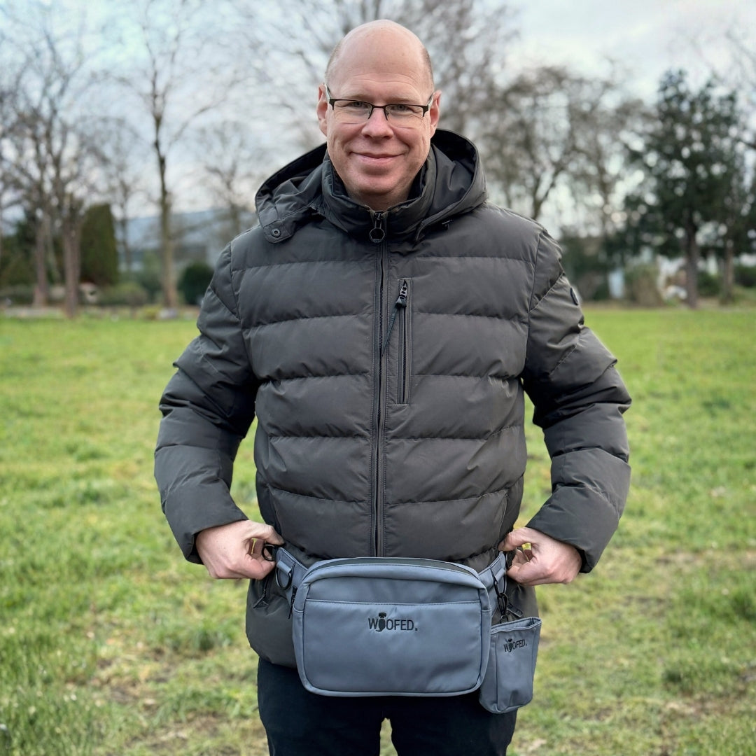 Graue Gassitasche/Bauchtasche frontal in Taillenhöhe, seitlicher Leckerlibeutel sichtbar, WOOFED-Logo, Mann auf grüner Wiese
