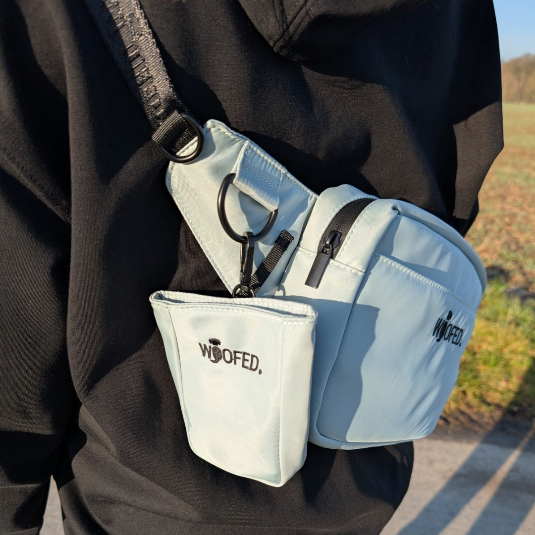 Salbeigrüne Gassitasche quer getragen, sichtbarer Reißverschluss, D‑Ring und abnehmbarer Leckerlibeutel mit WOOFED-Logo
