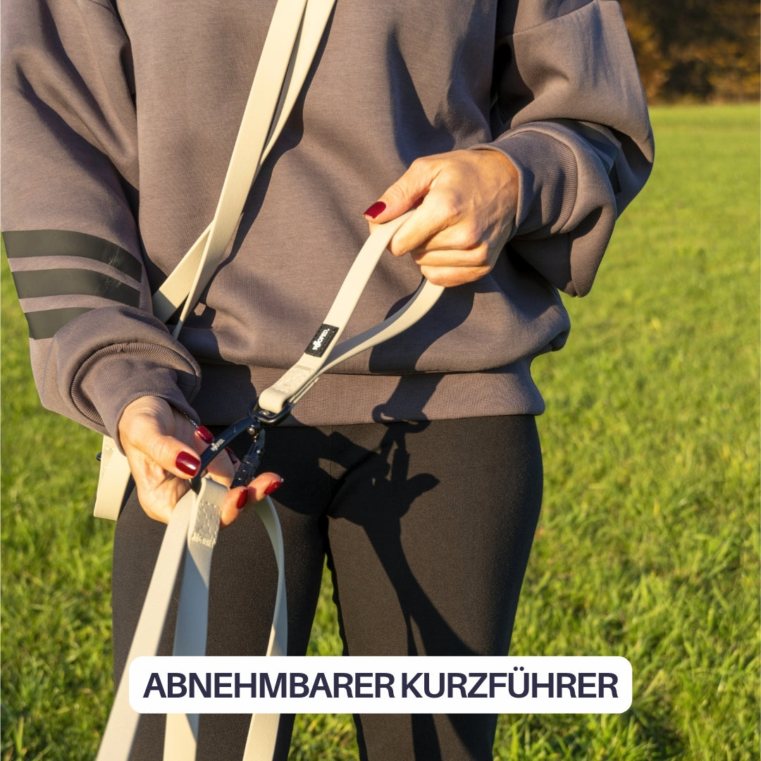 Beige Schleppleine mit abnehmbarem Kurzführer und schwarzem Karabiner, Nahaufnahme Hände an Hüfte vor Gras
