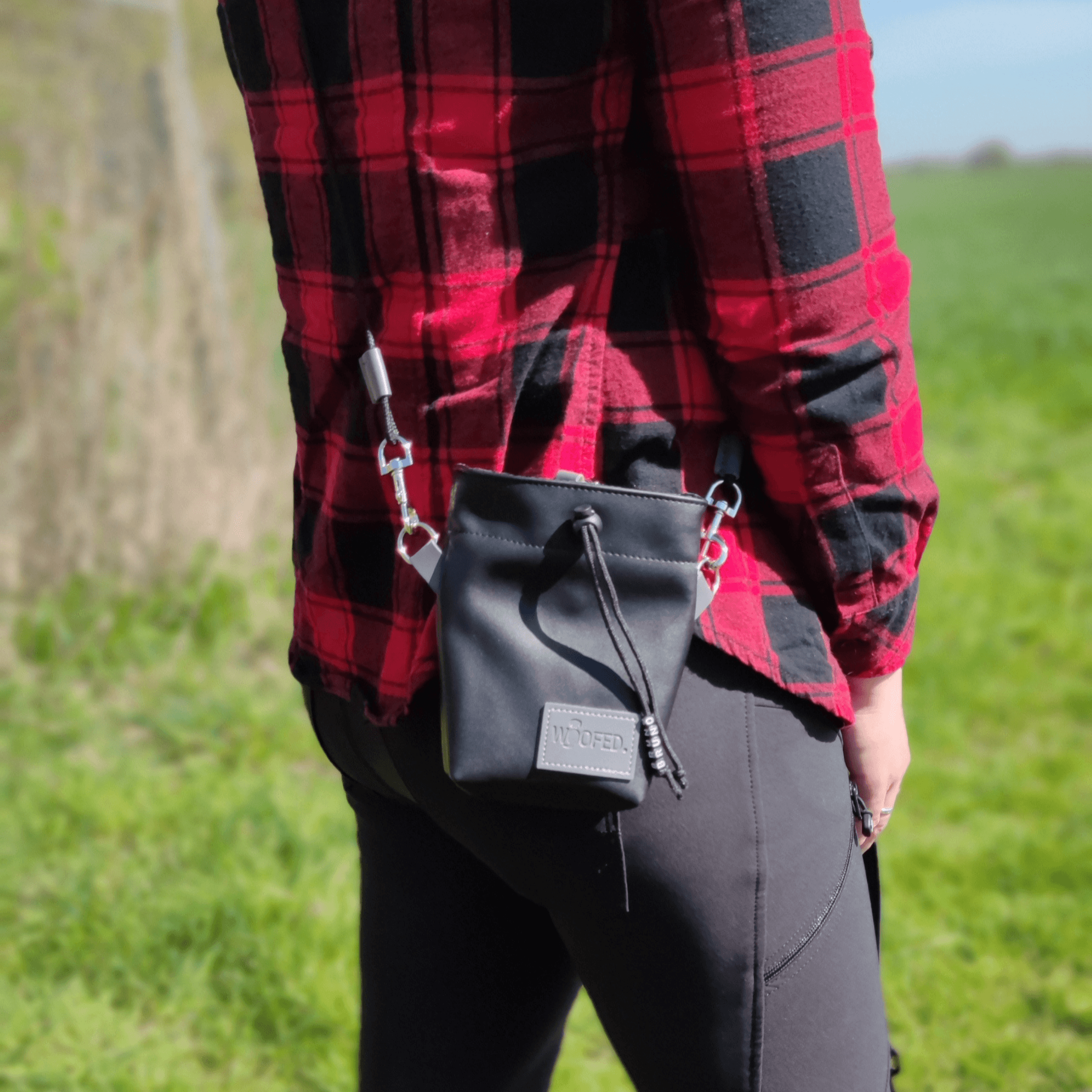 Schwarzer Leckerlibeutel Black Steel mit WOOFED-Prägung, Zugband und Metallclips, seitliche Ansicht an rotem kariertem Hemd